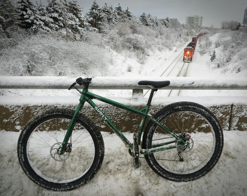 Left side view of a green Surly Krampus bike, parked in a snowbank on a bridge, with a train on tracks below