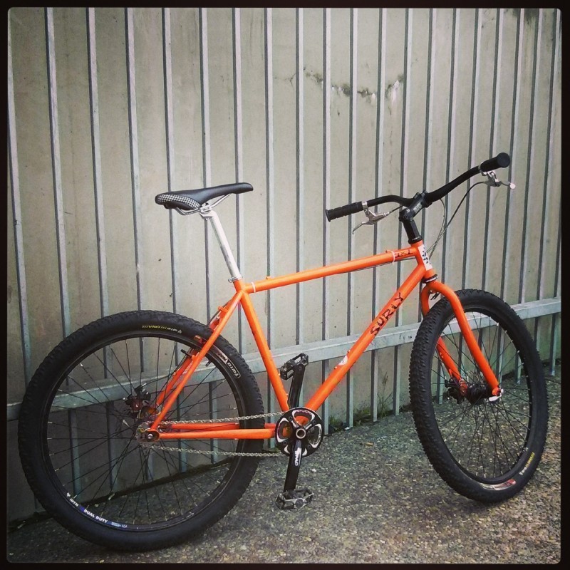 Right side view of an orange Surly 1x1 bike, leaning on a white, steel rod fence with a white wall behind it