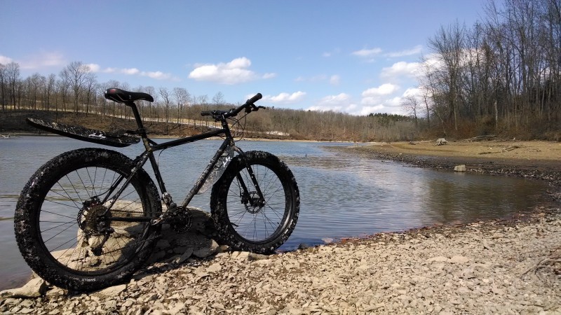 Right side view of a black Surly fat bike, parked along the rocky bank of a river, with bare trees on the sides