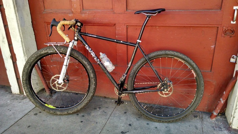 Left side view of a black Surly Karate Monkey bike, parked on the outside of a red, wooden garage door