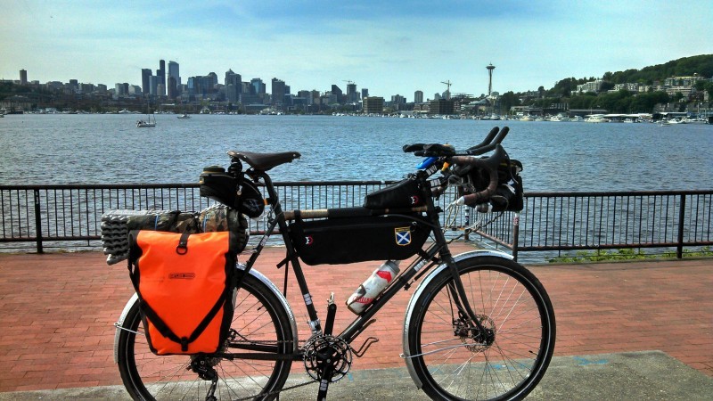 Right side view of a Surly bike loaded with gear, parked on a brick boardwalk, with a bay and city in the background