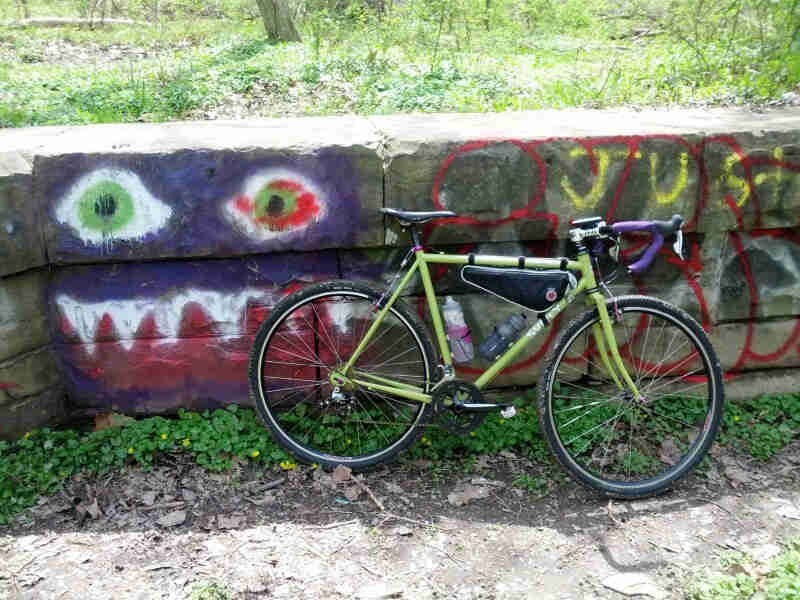 Right side view of a lime Surly bike, leaning against a stone block wall with graffiti, and grass above it
