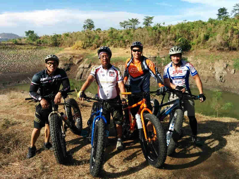 Front view of four cyclists, posing with their Surly fat bikes, with a retaining pond and green bushes behind them