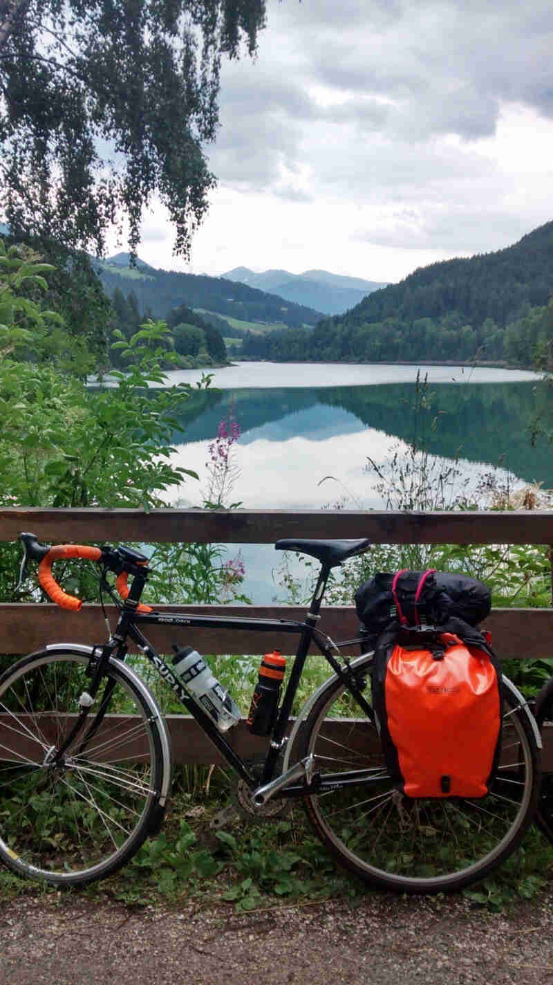 Left side view of a black Surly Cross Check bike, leaning on a wood fence, with a lake, trees and mountains behind it