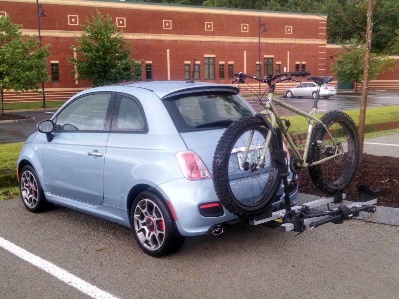 Left side view of a Surly fat bike, mounted to a rack on the back of a car, in a parking spot that's facing a building