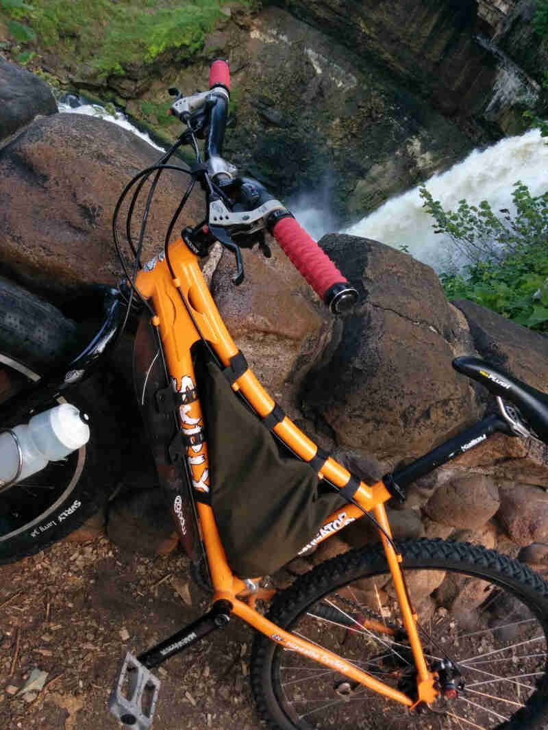 Left side view of an orange Surly Instigator bike, leaning on a rock wall, with stone cliffs and a waterfall behind it 