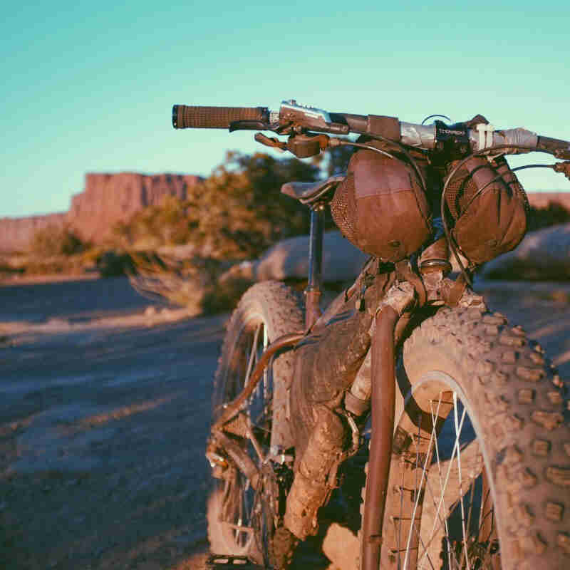 Front view of a muddy fat bike, with a desert field with bushes and buttes in the background