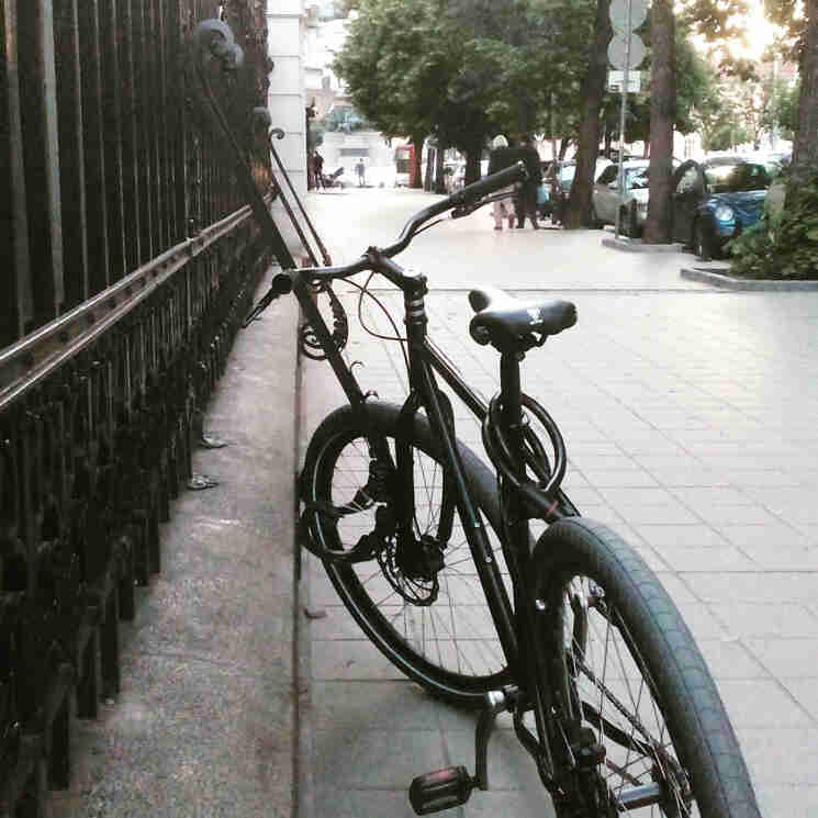Rear view of a black bike, facing down a sidewalk in a city, parked along an iron fence