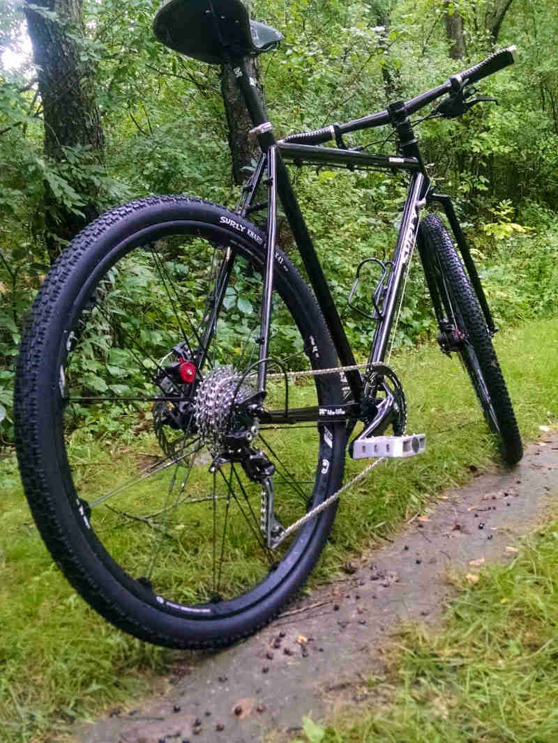 Rear, right side view of a black Surly bike, parked on the side of a grassy trail in the woods