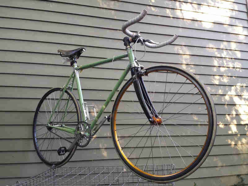 Right side view of a green bike, parked on top of a wire dog kennel, with the back wheel against the wall of a house