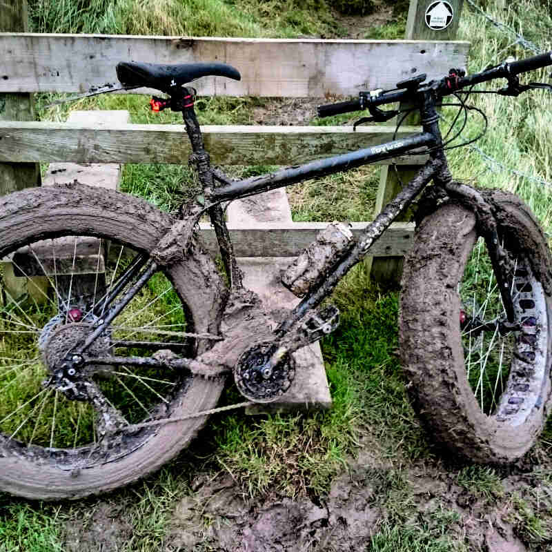 Right side view of a black Surly fat bike, caked with mud, standing in grass and leaning of a wood fence
