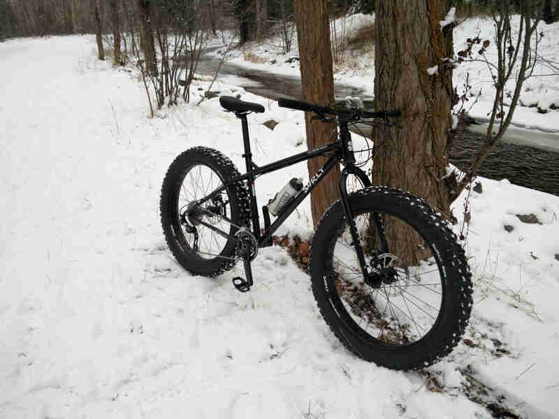 Front, right side view of a black Surly Ice Cream Truck fat bike, parked on snow against a tree, with a stream behind