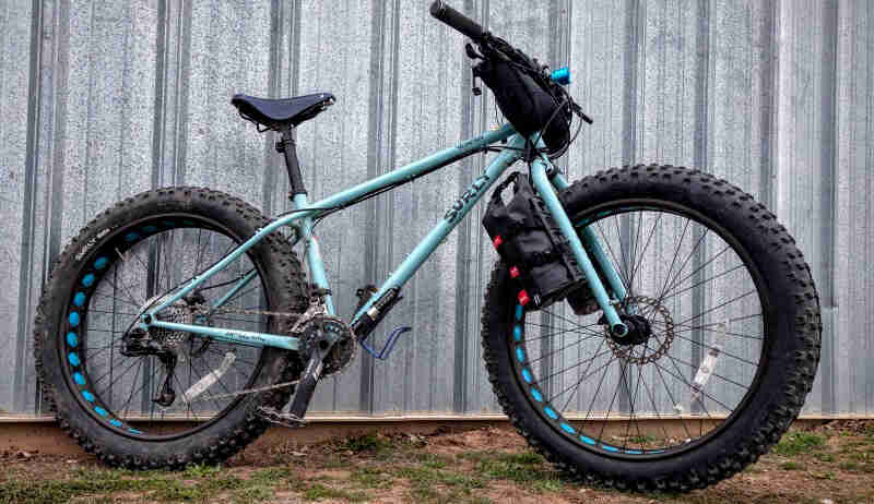Right side view of a Surly Ice Cream Truck fat bike, turquoise, parked on dirt and grass, in front of a steel shed wall