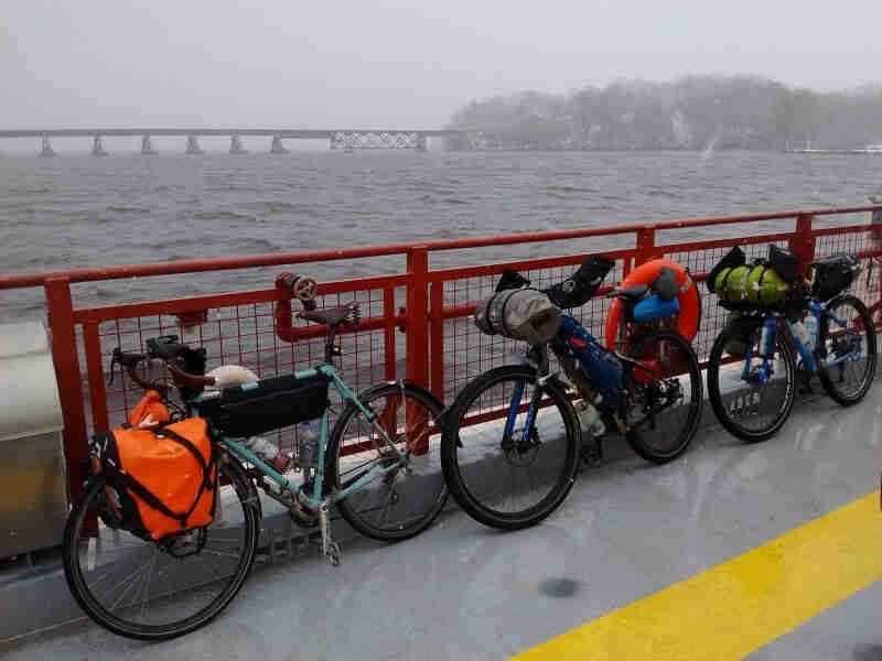 Left side view of 3 bikes, loaded with gear, parked in single file along a railing, in front of a lake on a foggy day