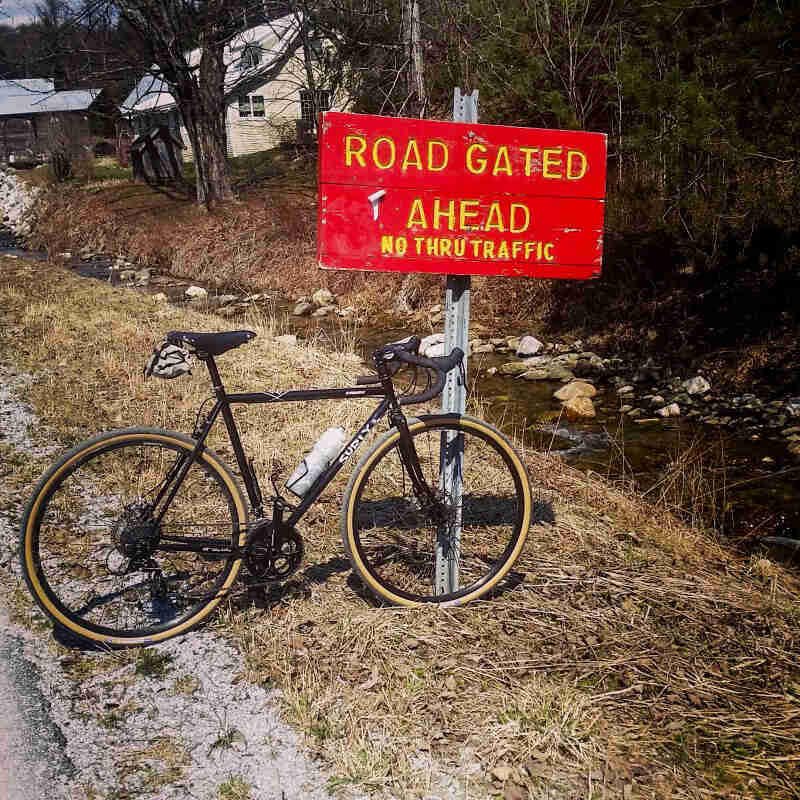 Right side view of a Surly bike, parked on the side of a road, in front of a sign, and a stream and houses in the background