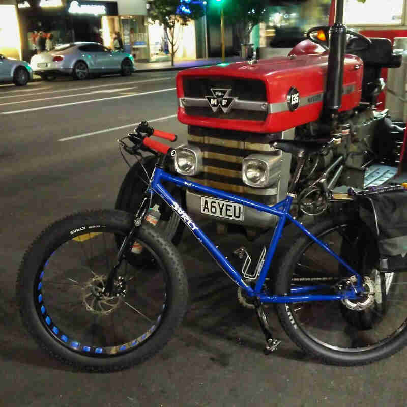 Left side view of a blue Surly Troll bike, parked on a street, against the front of a Massey Ferguson tractor, at night