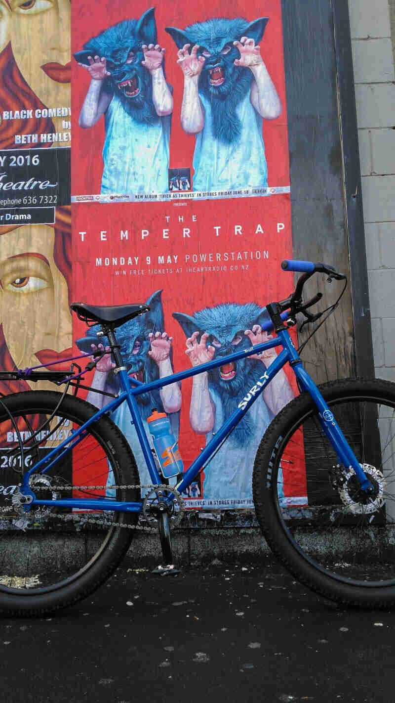 Right side view of a Surly Troll bike, blue, on a sidewalk, leaning on a wall with, The Temper Trap, theater ad posters