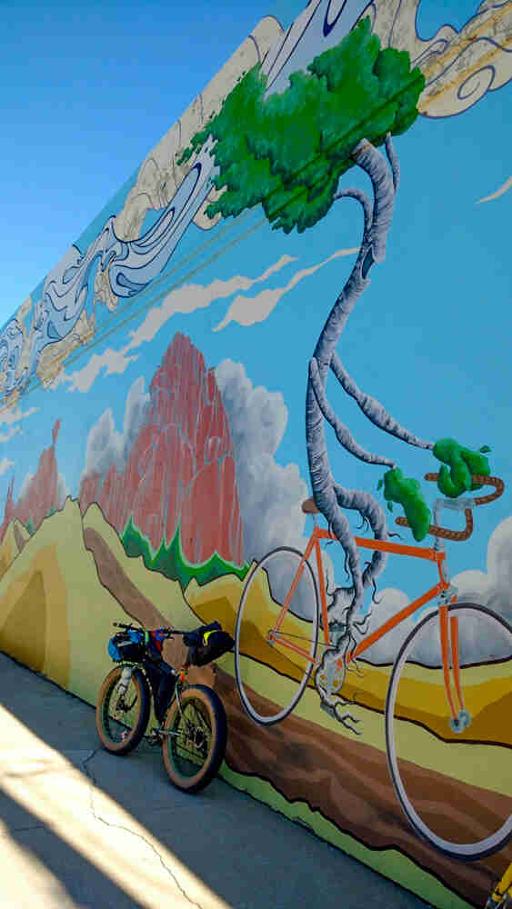 Rear view of a Surly fat bike, loaded with gear, parked on pavement, leaning on a wall painted with a mural