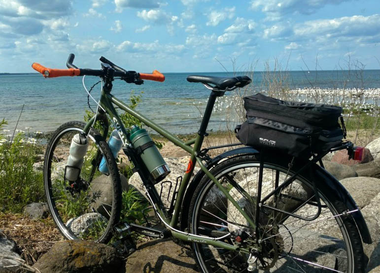 Rear left view of a Surly bike, olive, parked on a rocky shore, facing a lake