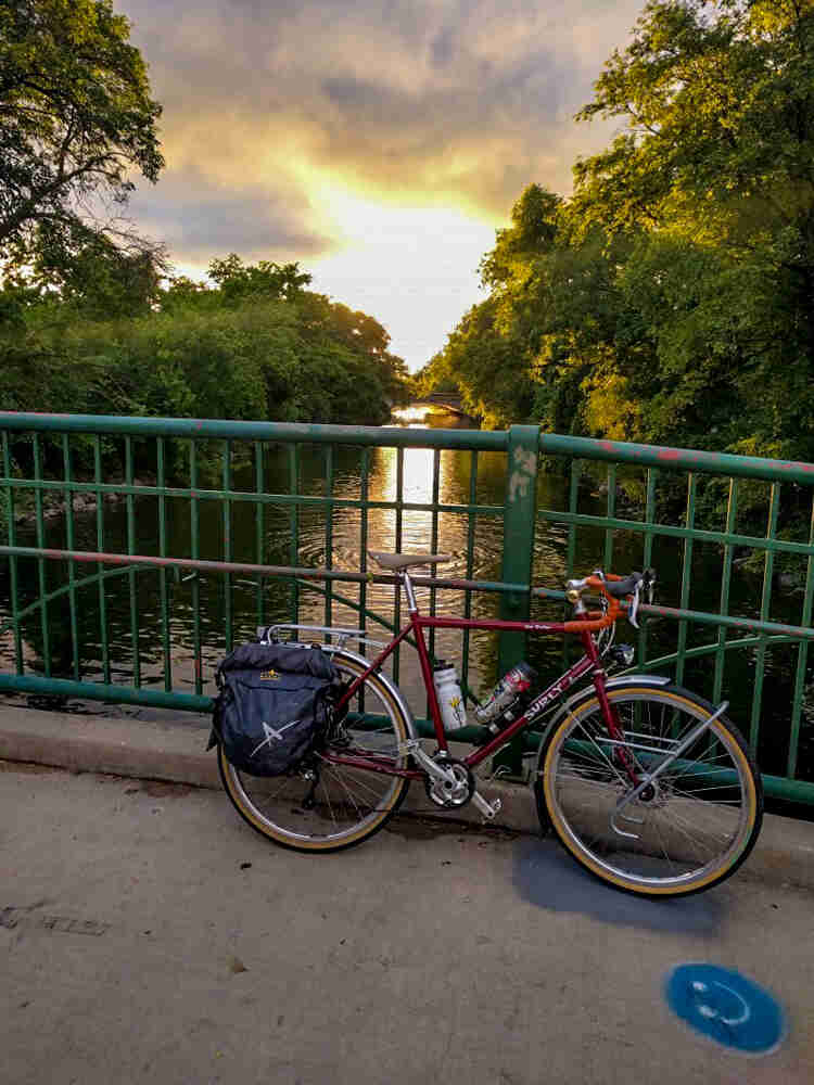 A Surly Disc Trucker bike - red - parked along the rail of a bridge - with a river below and trees on both sides