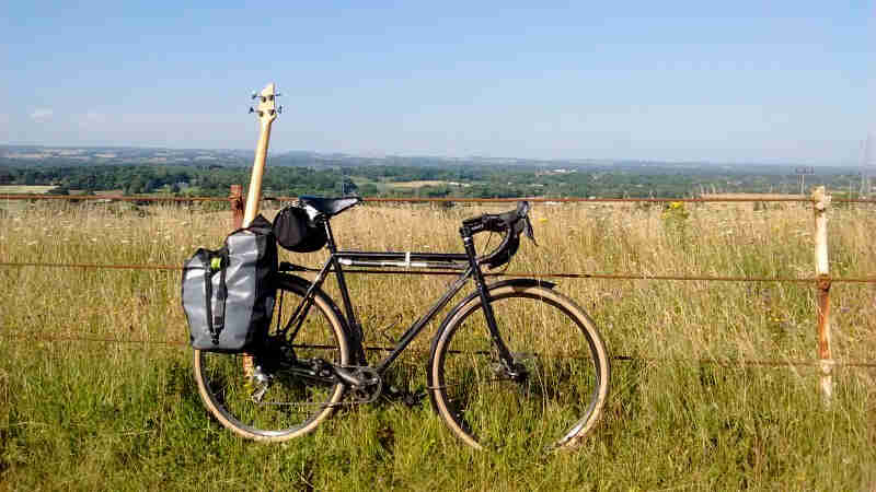 Right side view of a black Surly bike with a guitar on back, parked in tall grass, in front of a barbed wire fence