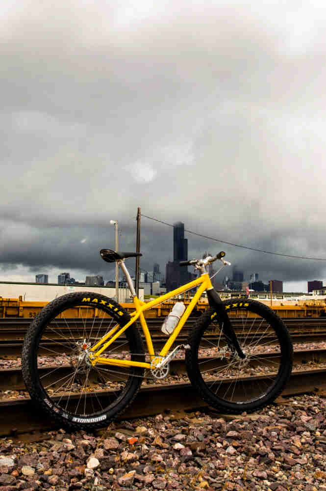 Right side view of a yellow Surly bike, next to tracks at a rail yard, with a city skyline and clouds in the background