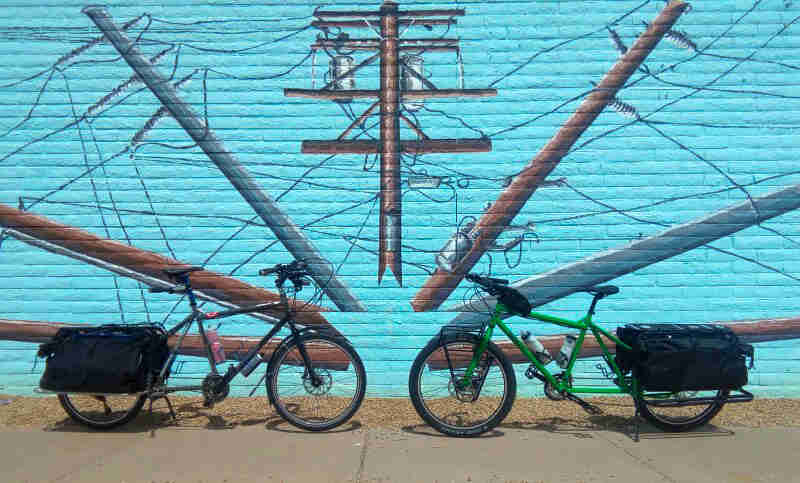 Side view of 2 Surly bikes facing each other, parked on a side walk in front of a brick wall with a mural painted on it