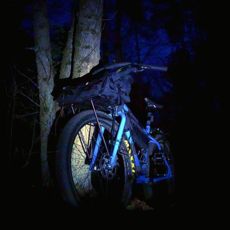 Front left side view of a Surly bike, blue, loaded with gear, leaning on a tree at night