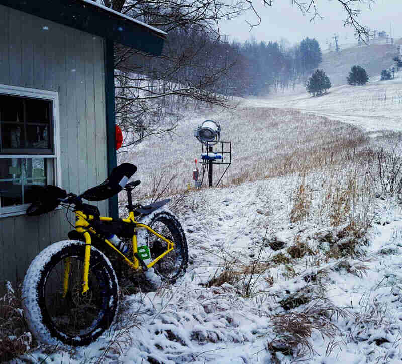 Front left side view of a Surly fat bike in the snow, leaning on a building, with trees and a hill in the background
