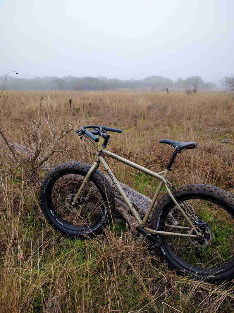 Left side view of a Surly fat bike standing in a field of brush, with trees and fog in the background