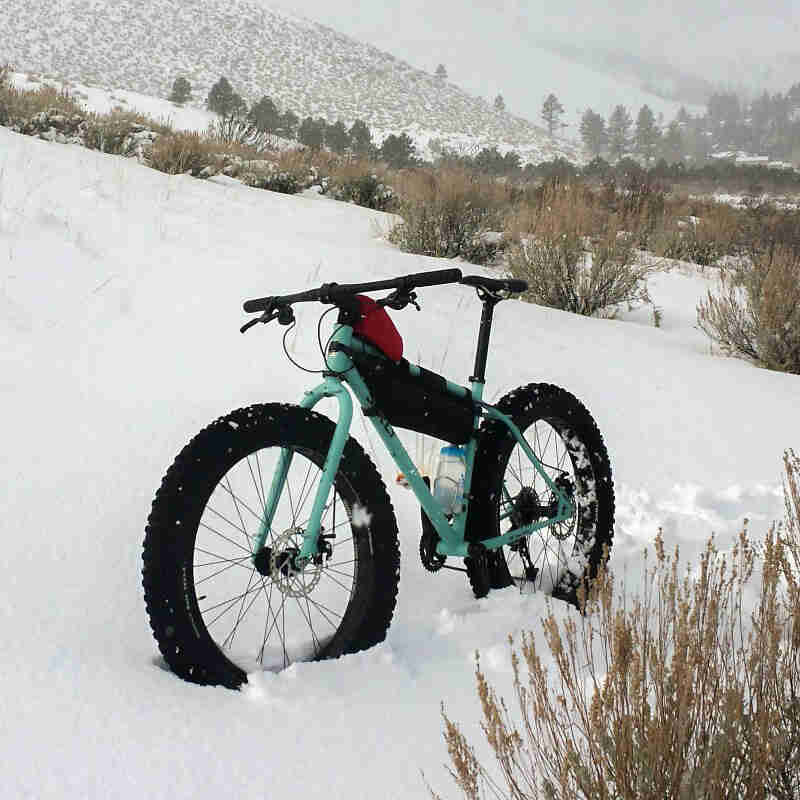 Front left view of a Surly fat bike, at the base of a snow covered hill, with trees and mountain in the background