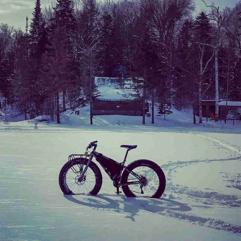 Left side view of a Surly fat bike standing in deep snow on a frozen lake, with a cabin in the background