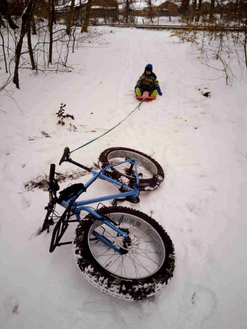 Front view of a Surly fat bike laying in the snow, with a child in a sled tied to the seat further behind in background