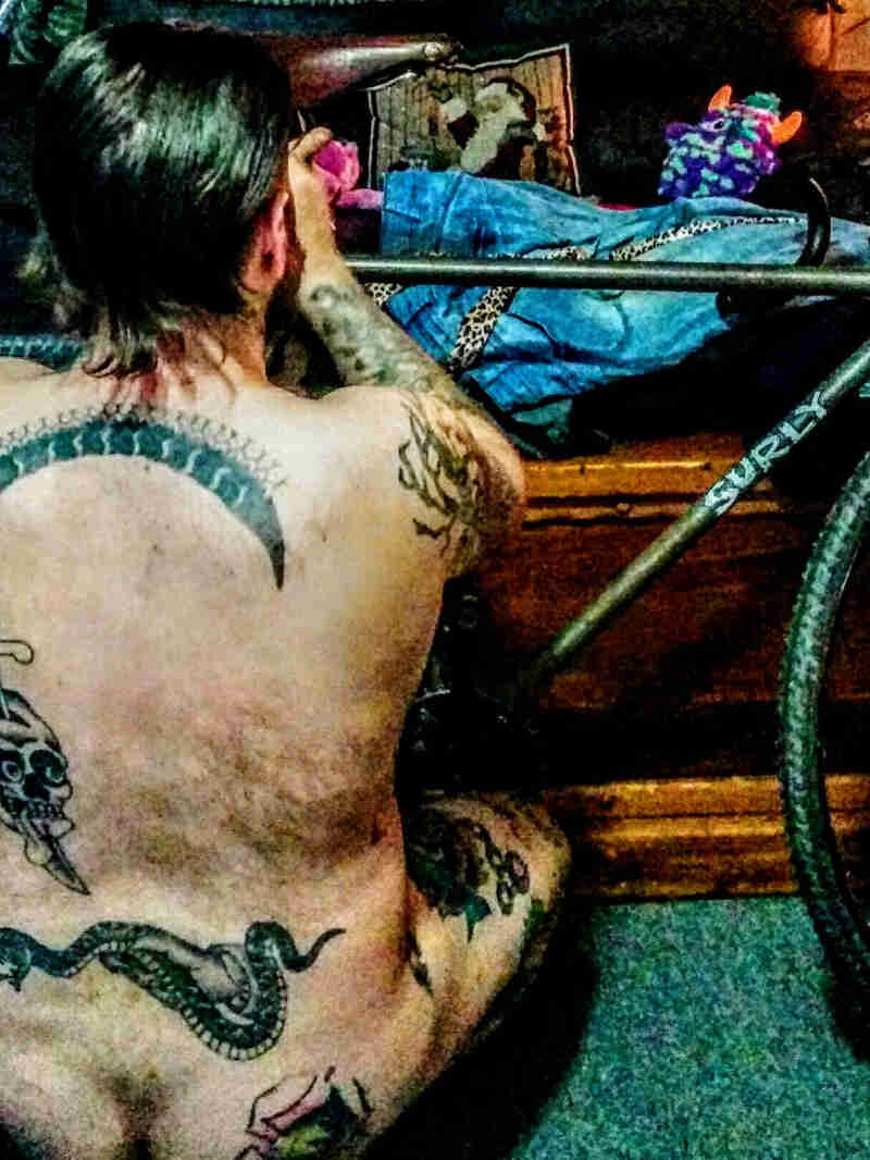 Rear of a person with no clothes and tattoos on their back, squatting down in front of bike adjusting the seat