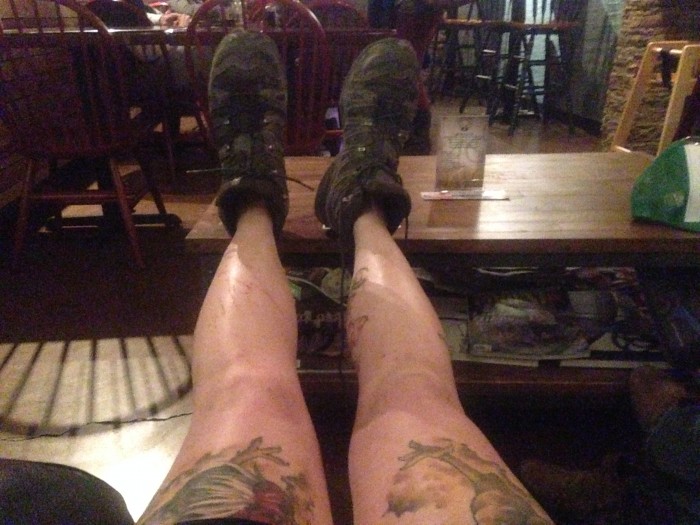 A person's legs with tattoos and wearing hiking boots, face straight away with their feet propped up on a coffee table