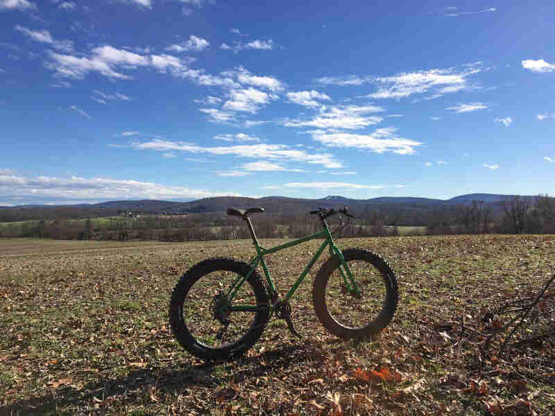 Right side view of a green fat bike, parked on leaves in a large grass field, with hills in the background