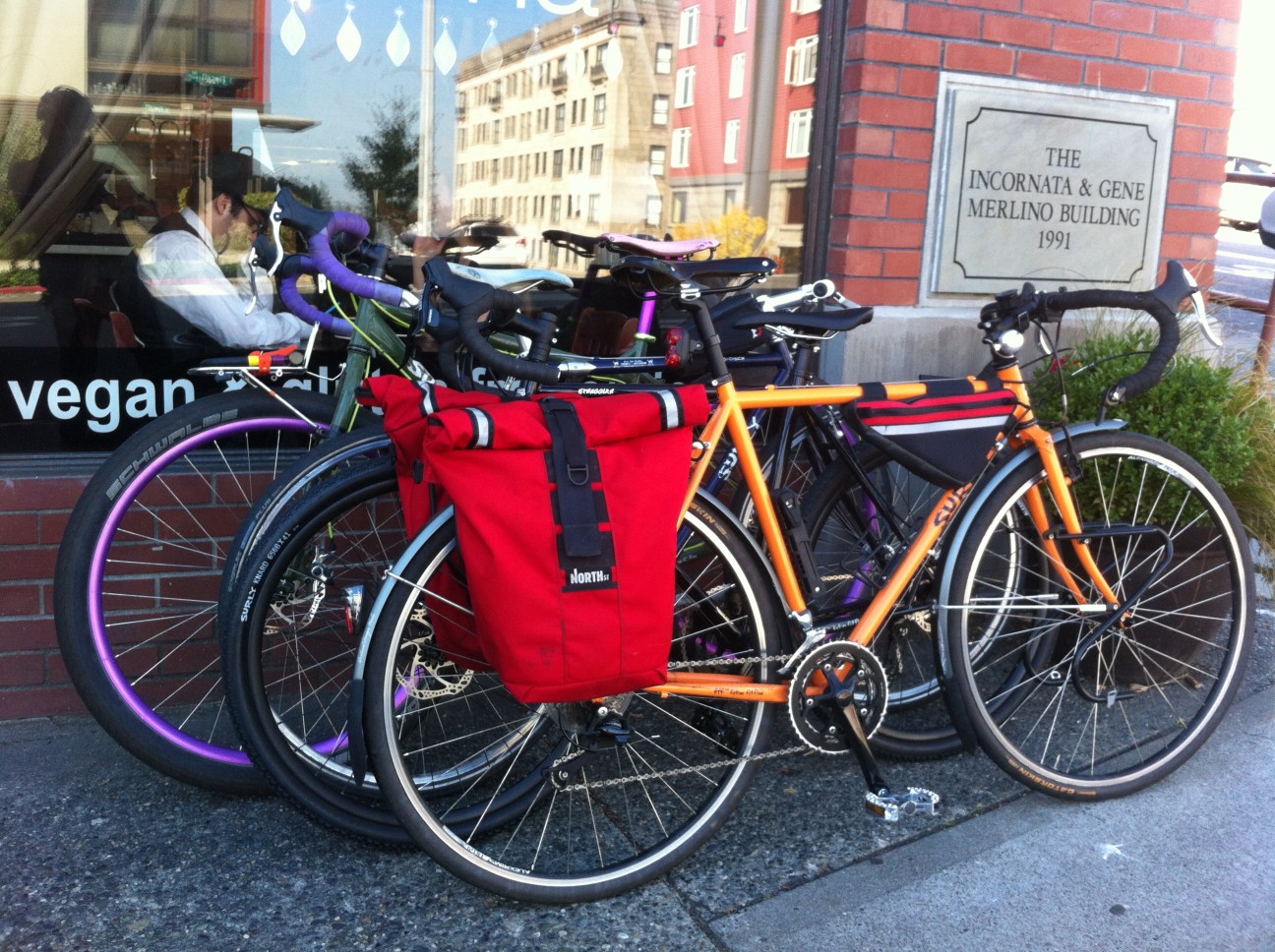 Right side view of Surly bikes, parked side by side, on a sidewalk in front of a window on a building