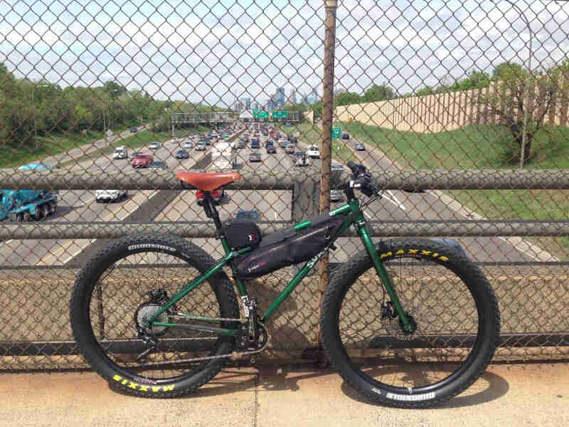 Right side view of a green Surly Krampus bike, parked along the rail, on a bridge over a freeway, with a city behind