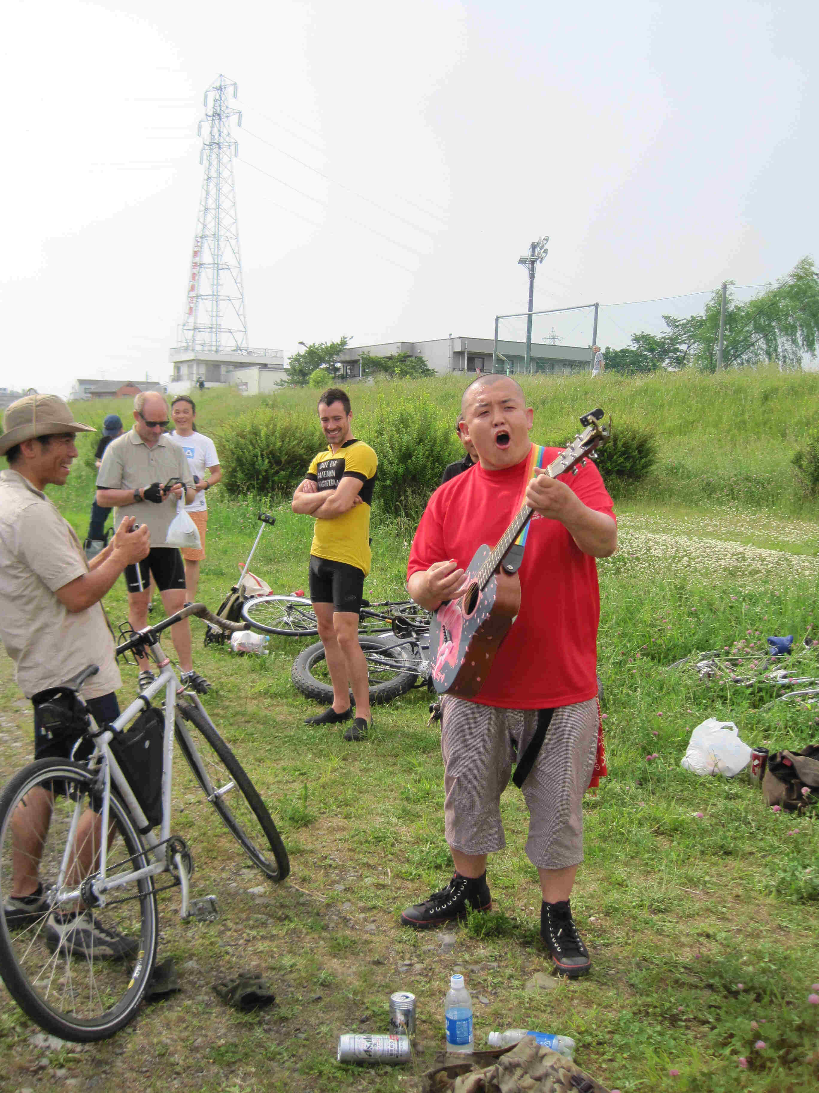 Front view of a person playing a guitar and singing, in a grass field, with bikes and cyclists around them