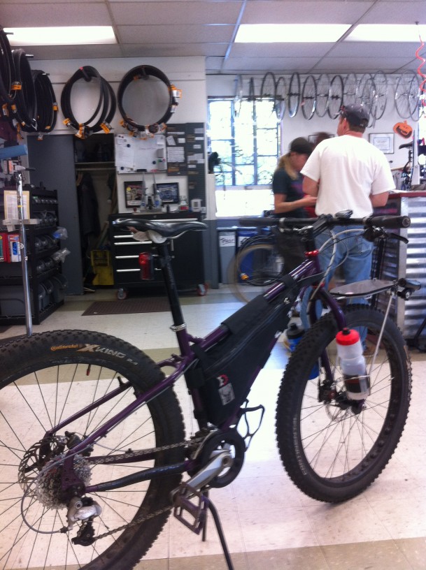 Rear, right side view of a purple Surly fat bike with a frame bag, with people in the background, in a bike shop