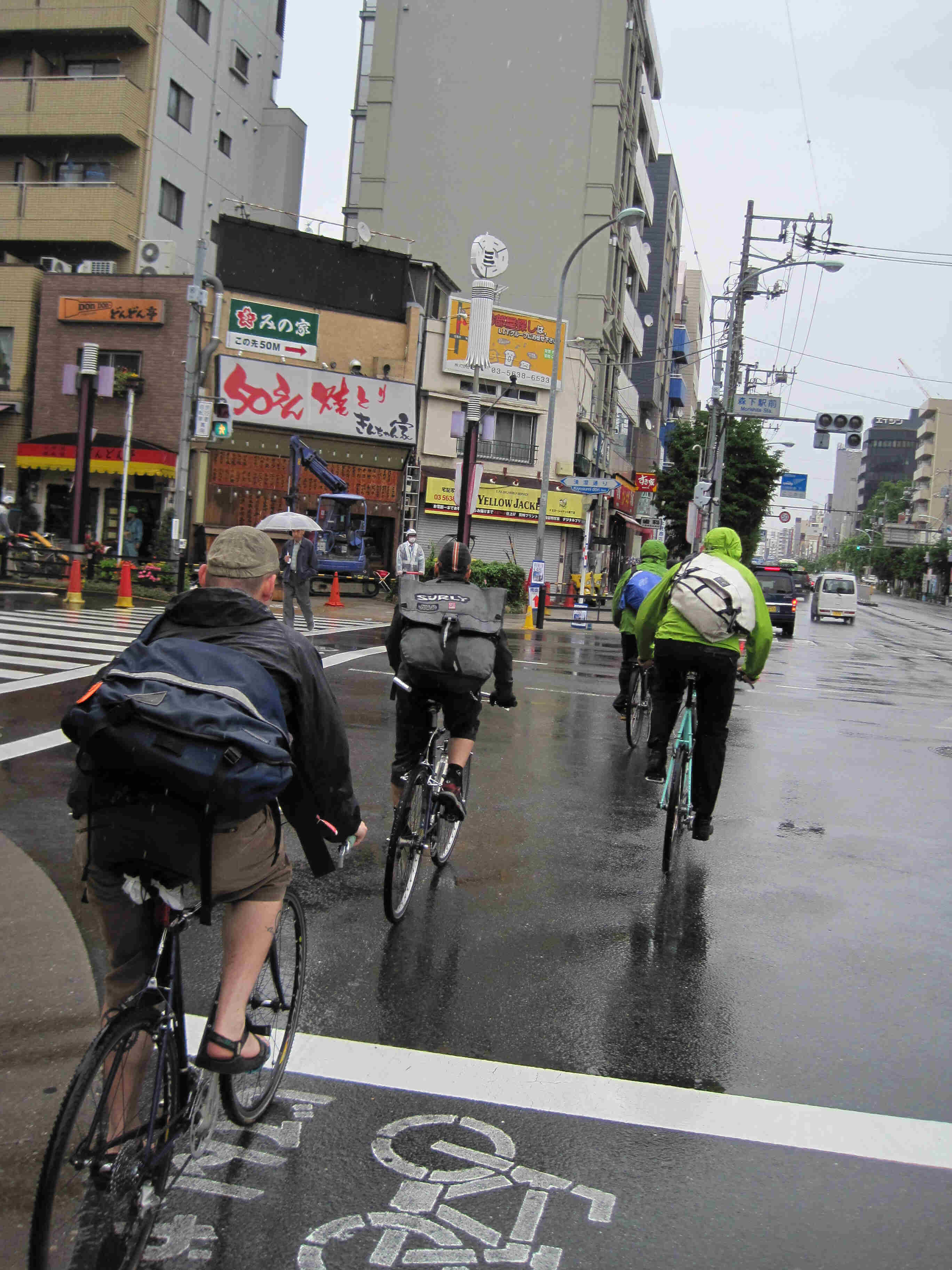 Rear view of 4 cyclists, riding their Surly bikes across a wet city street, with building on both sides