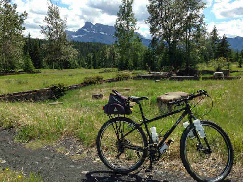 Right side view of a black Surly bike, parked on a trail, with a grass field, trees and mountains in the background