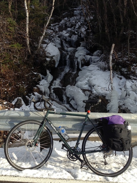 Left side view of a green Surly Disc Trucker bike, leaning on a guardrail in snow, with a frozen waterfall behind