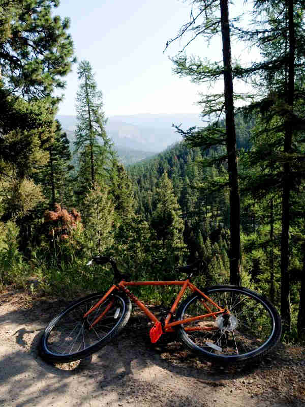 Left side view of an orange Surly bike lying on it's side, on a cliff edge with tree covered mountains in background