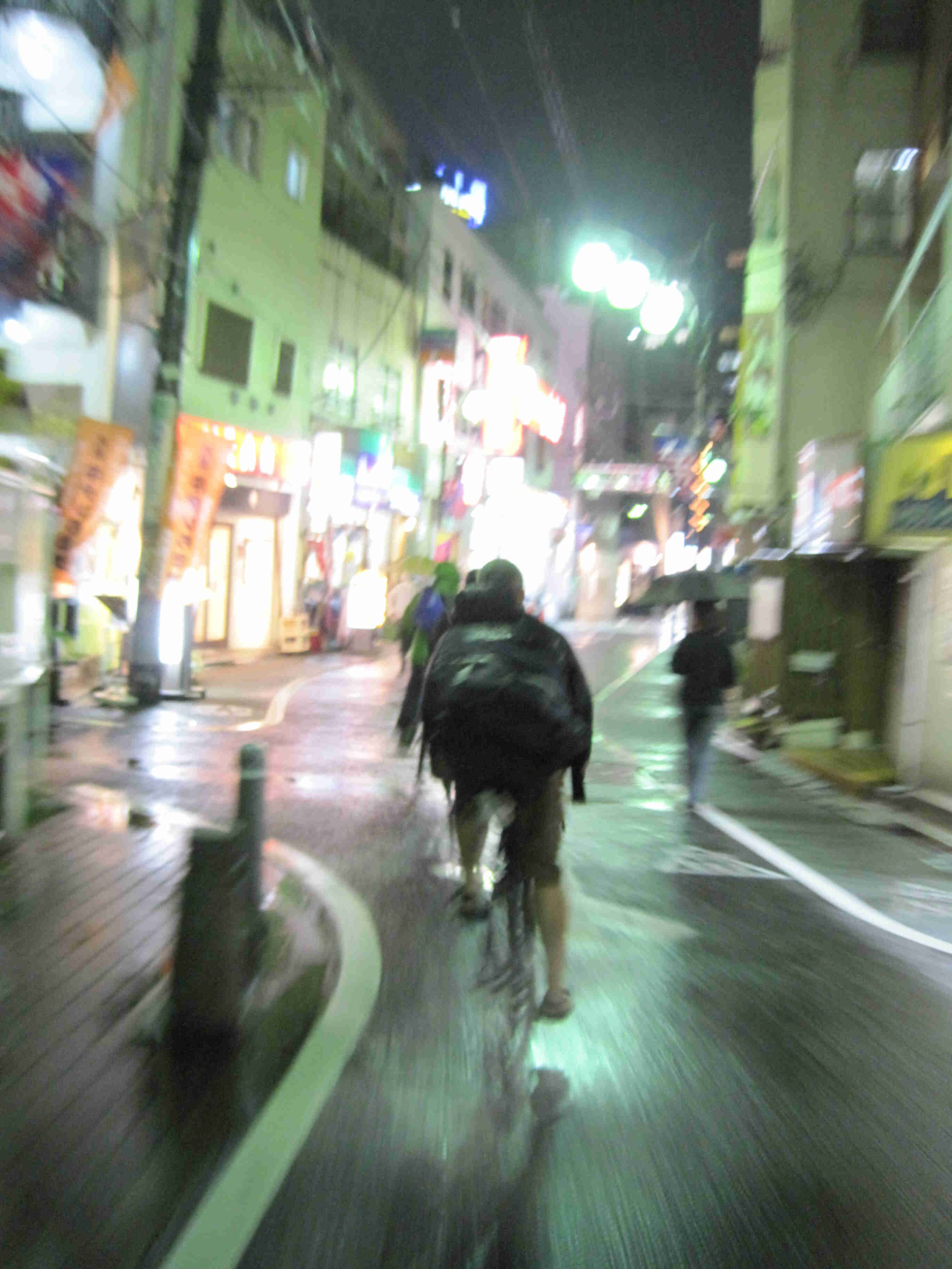 Rear view of cyclists, riding in single file at night, straight away on a narrow city street, with buildings on the side