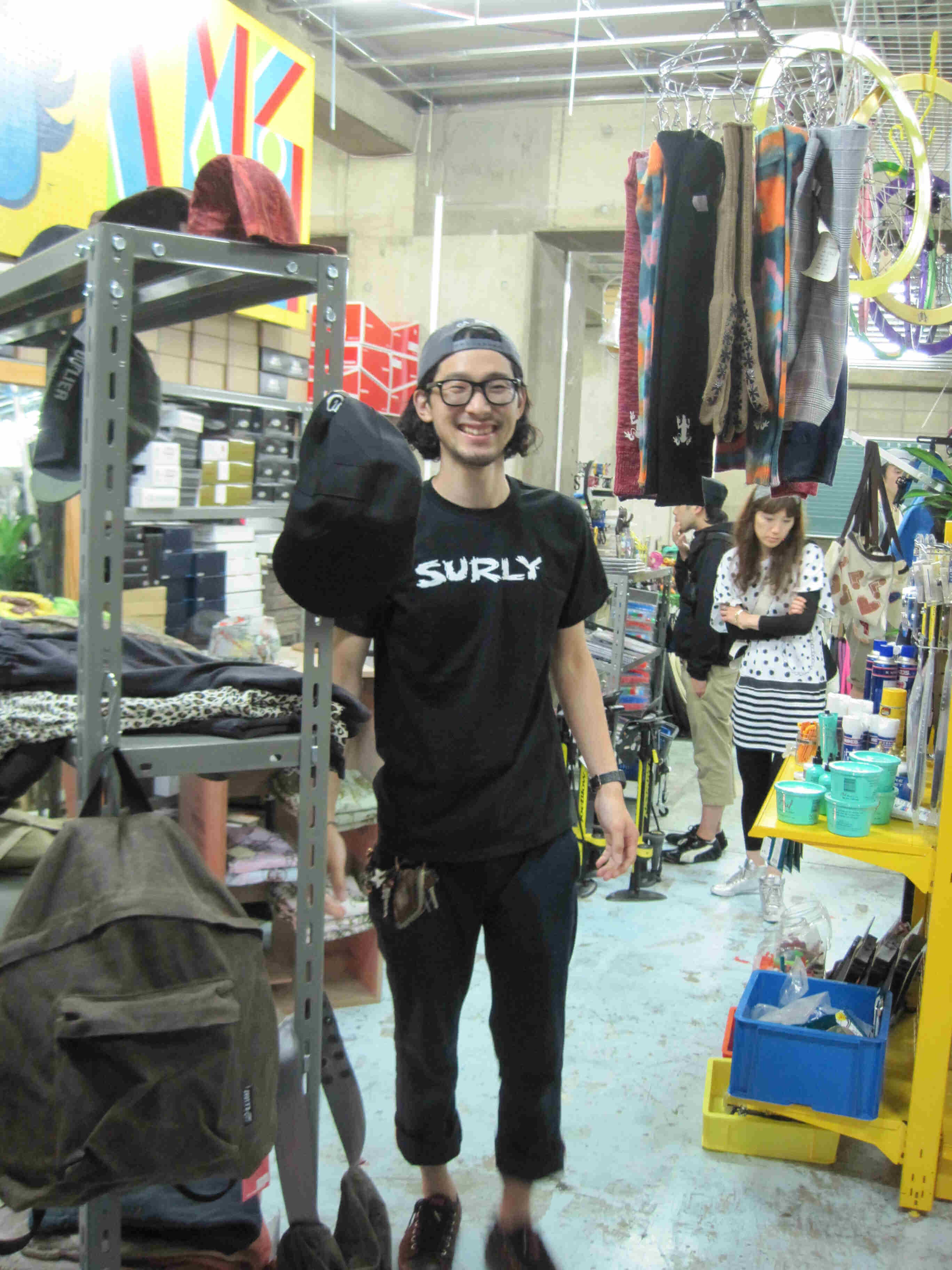 Front view of a person, wearing a black Surly Bikes t-shirt, standing in a bike shop, next to a rack with clothes on it