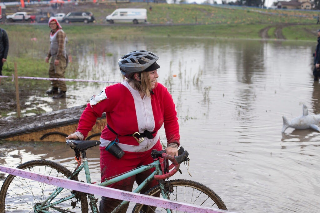 Cyclist Marley happily wearing a santa suit and bike helmet walking a bike across a muddy pond