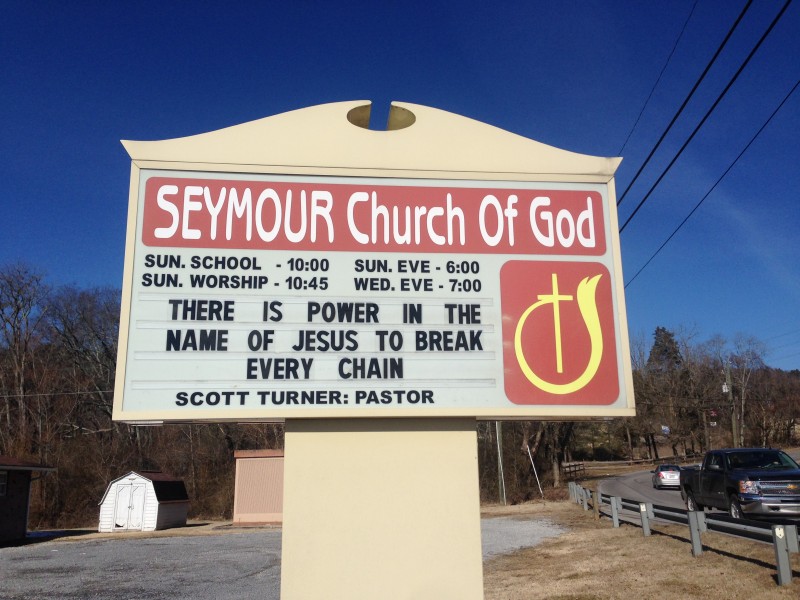 A Seymour Church Of God sign that shows, The is power in the name of Jesus to break every chain, near a parking lot