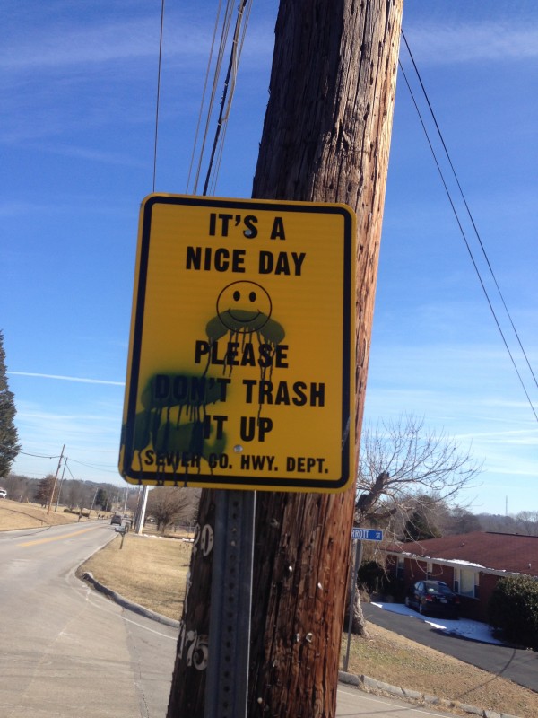 Front view of a yellow street sign with black text, in front of a telephone pole, on the side of a paved road