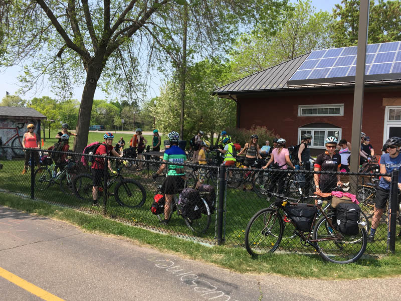 A group of cyclists gathered on a grassy, fenced in area, next to a red building with solar panels on the roof